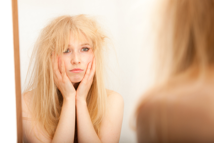 A young blonde woman, head in hands, fed up with her hair as she looks into the mirror.